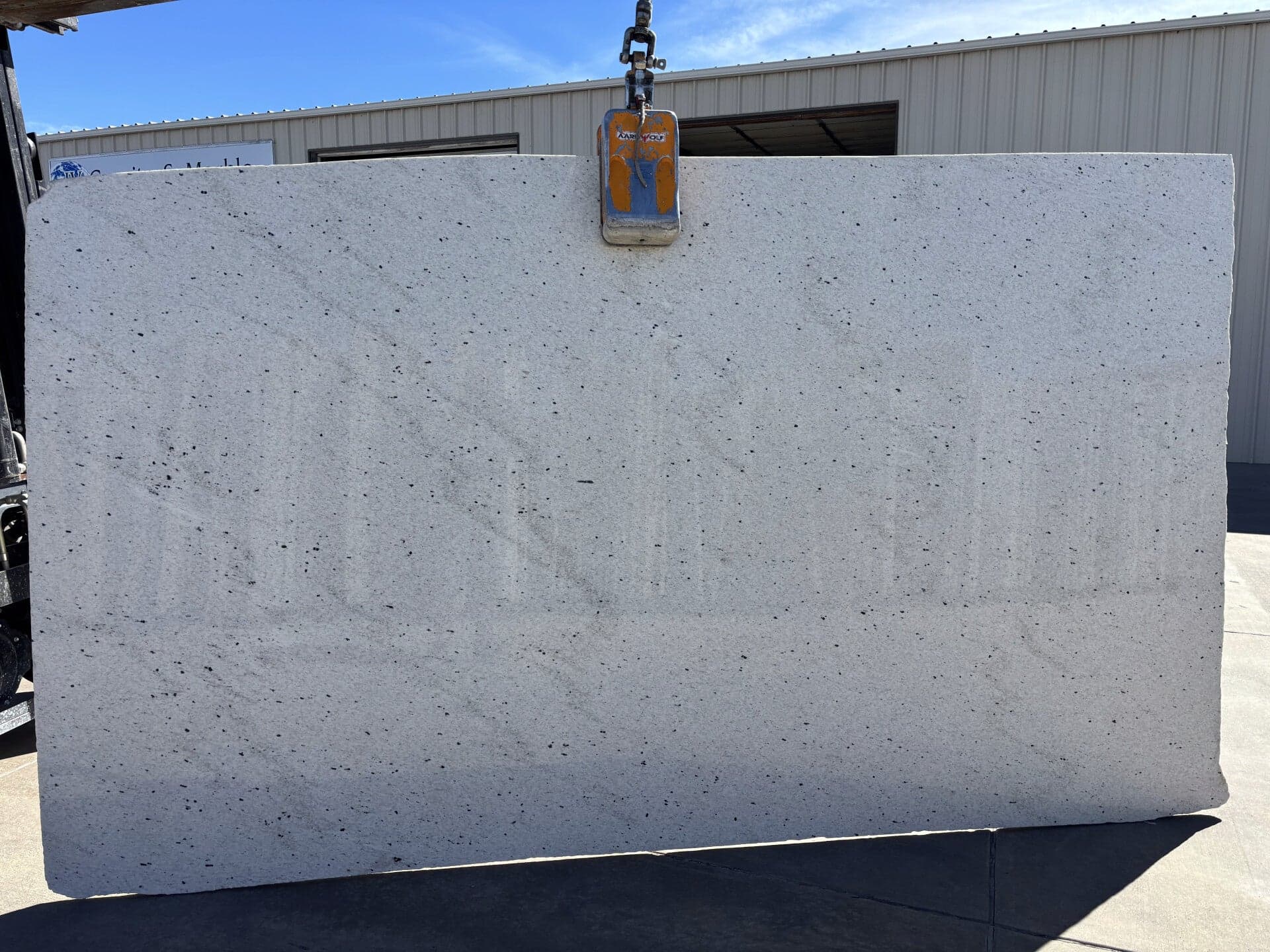 A white granite slab hanging from a lifting clamp outside the I.V. Granite & Marble shop